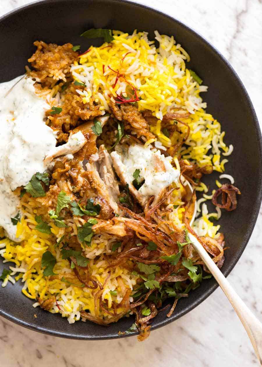 Chicken Biryani in a rustic black bowl with yellow saffron rice, garnished with crispy fried onions, coriander and minted yoghurt
